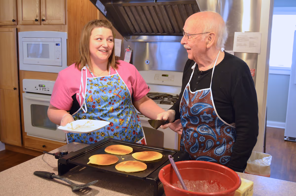 An elderly man and a younger woman wearing aprons are cooking pancakes on a griddle in a kitchen. The woman is holding a plate and smiling at the man, who is holding a spatula. The kitchen has wooden cabinets, a microwave, an oven, and a stove with a stainless steel hood.