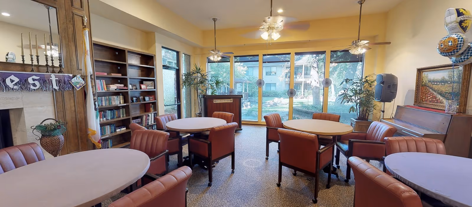 Community activity room with round tables and upholstered chairs, bookshelves, a piano, and large windows overlooking a garden.