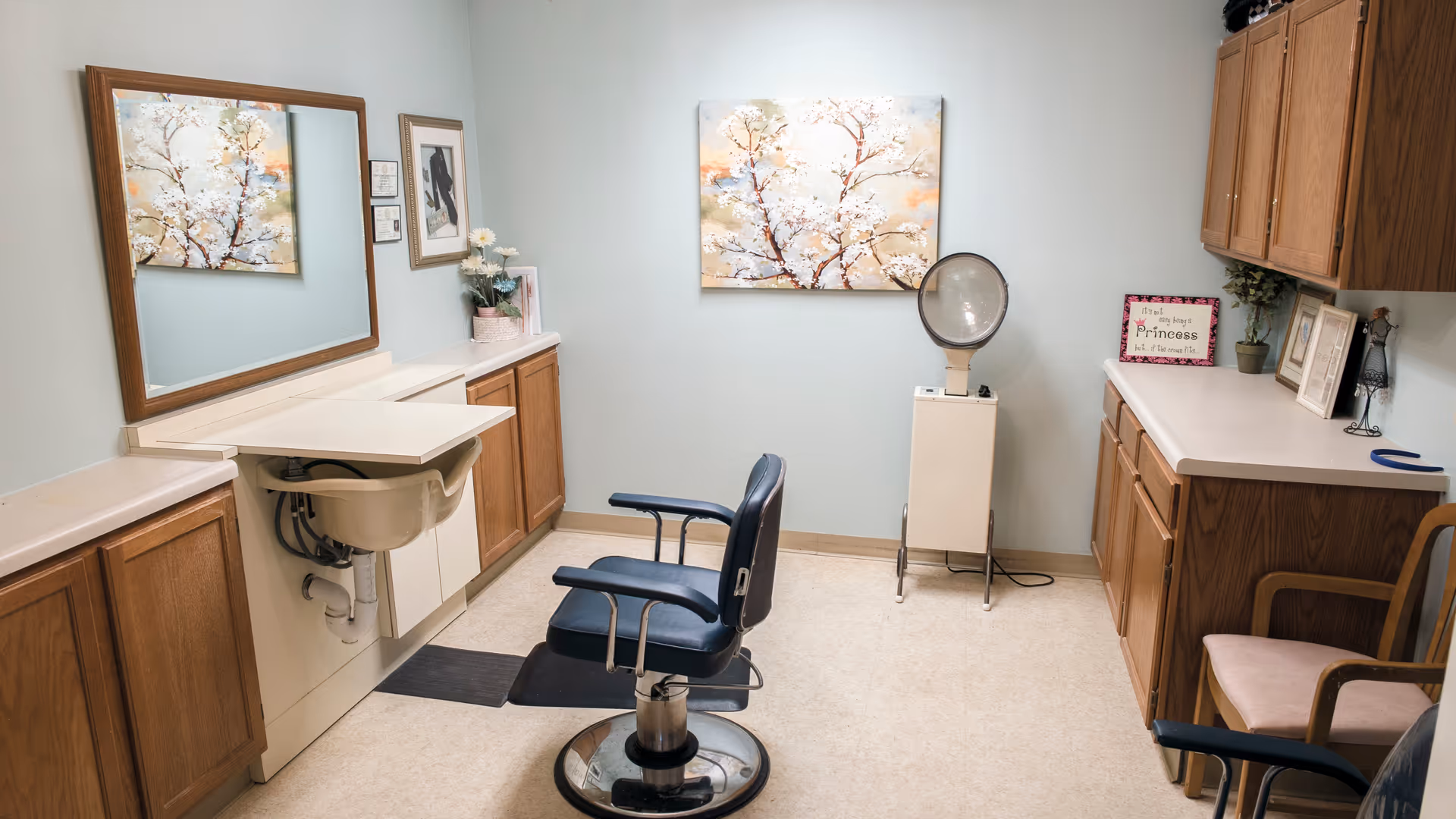 A small salon room with a black salon chair in front of a large mirror mounted on the wall. There is a sink below the mirror and wooden cabinets on both sides of the room. On the right side, there is a vintage hair dryer and decorative items including framed pictures and a sign that reads 'It's not easy being a Princess but... it's the crown I fit.' The walls are light blue and there is a painting of white blossoms on a tree hanging on the wall.