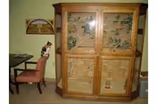 A large wooden enclosure with glass doors housing a multi-level habitat for small animals or birds. A calico cat is sitting on a pink chair next to the enclosure. The room has light-colored walls and a table partially visible on the left side.
