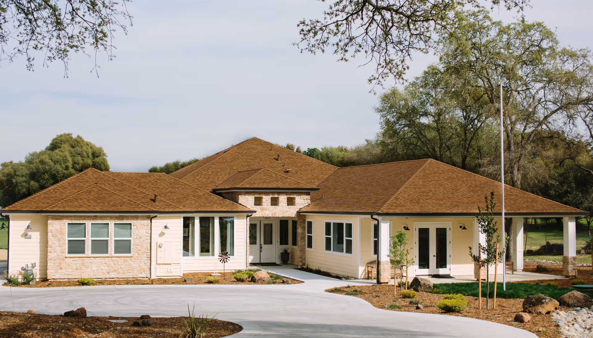 Front exterior view of a single-story building with a brown shingle roof, light yellow siding, and stone accents around the entrance. The building is surrounded by a landscaped area with small trees, shrubs, and a curved concrete driveway. Large trees and a clear sky are visible in the background.