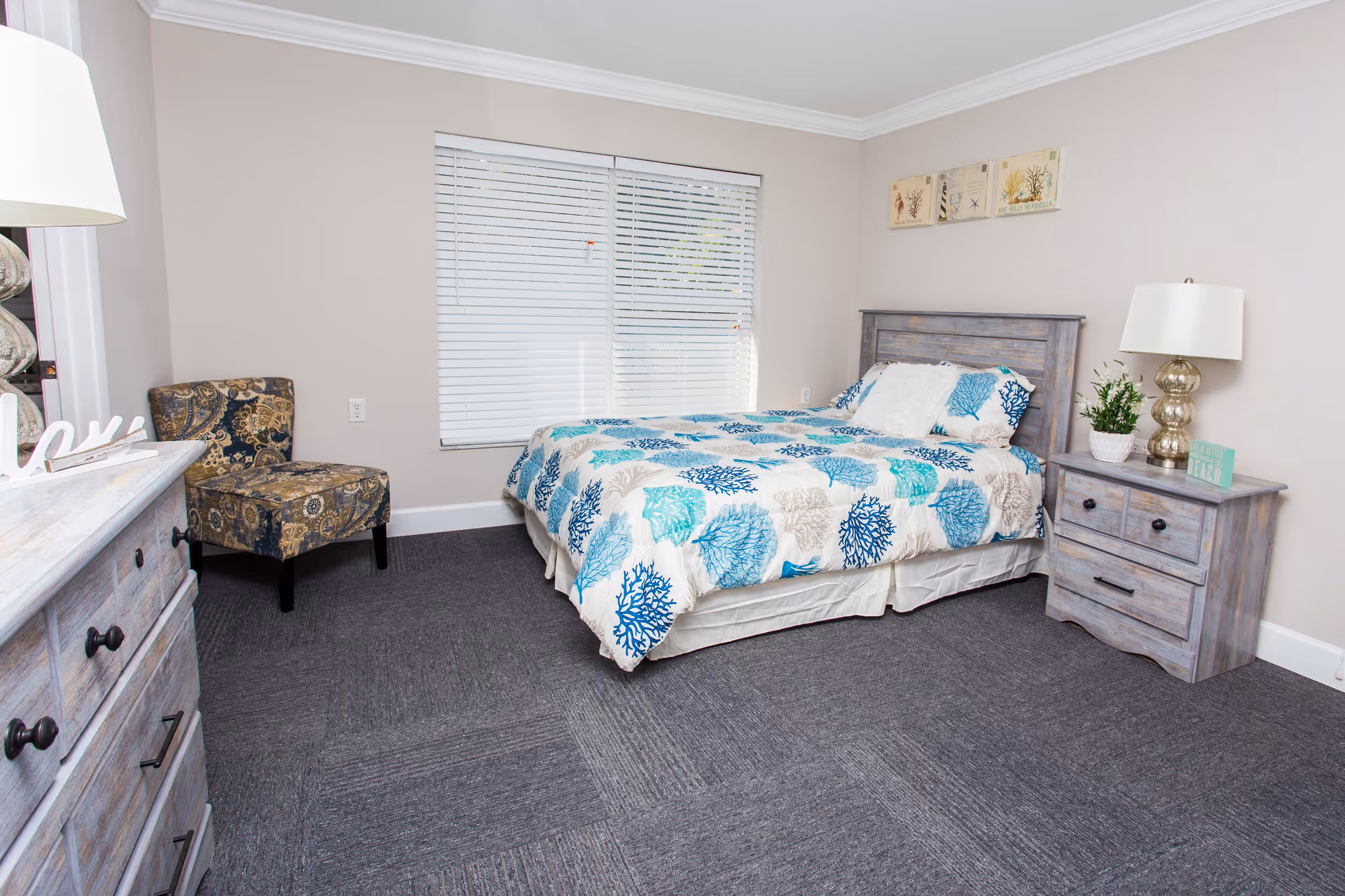 A bright bedroom with a bed covered in a blue and beige coral-patterned comforter, a wooden headboard, a matching wooden nightstand with a lamp and a plant, a patterned upholstered chair in the corner, a wooden dresser with a decorative 'love' sign, and a window with white blinds.