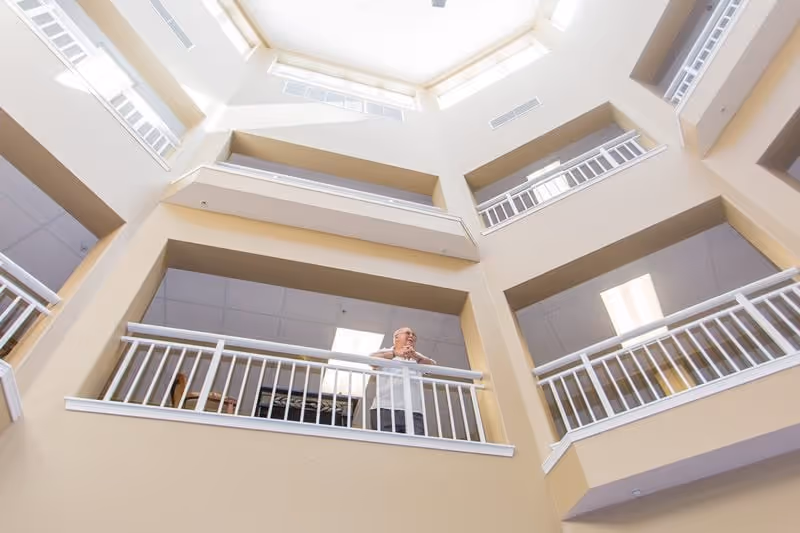 An elderly man standing on an indoor balcony railing inside a multi-story building with beige walls and white railings, looking outward. The ceiling above is a skylight allowing natural light to illuminate the space.