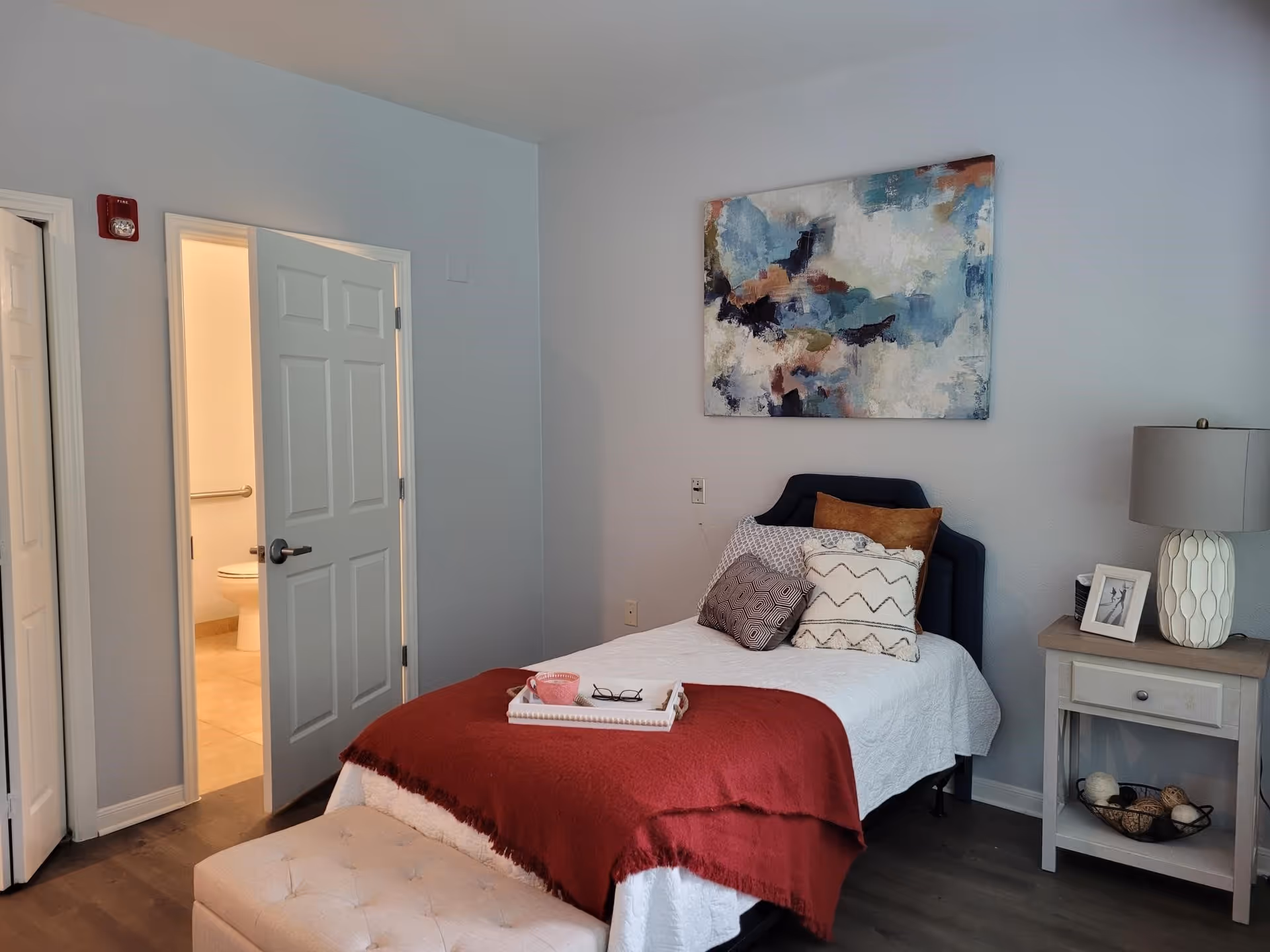 A tidy single bedroom with a made bed, bedside table and lamp, abstract wall art, and an open door to a bathroom.
