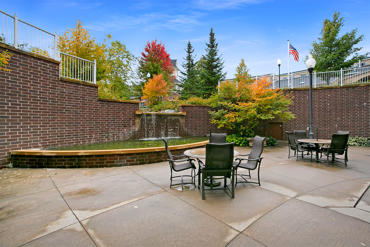Outdoor courtyard with patio tables and chairs, a brick water feature with a small waterfall, and trees with autumn foliage.