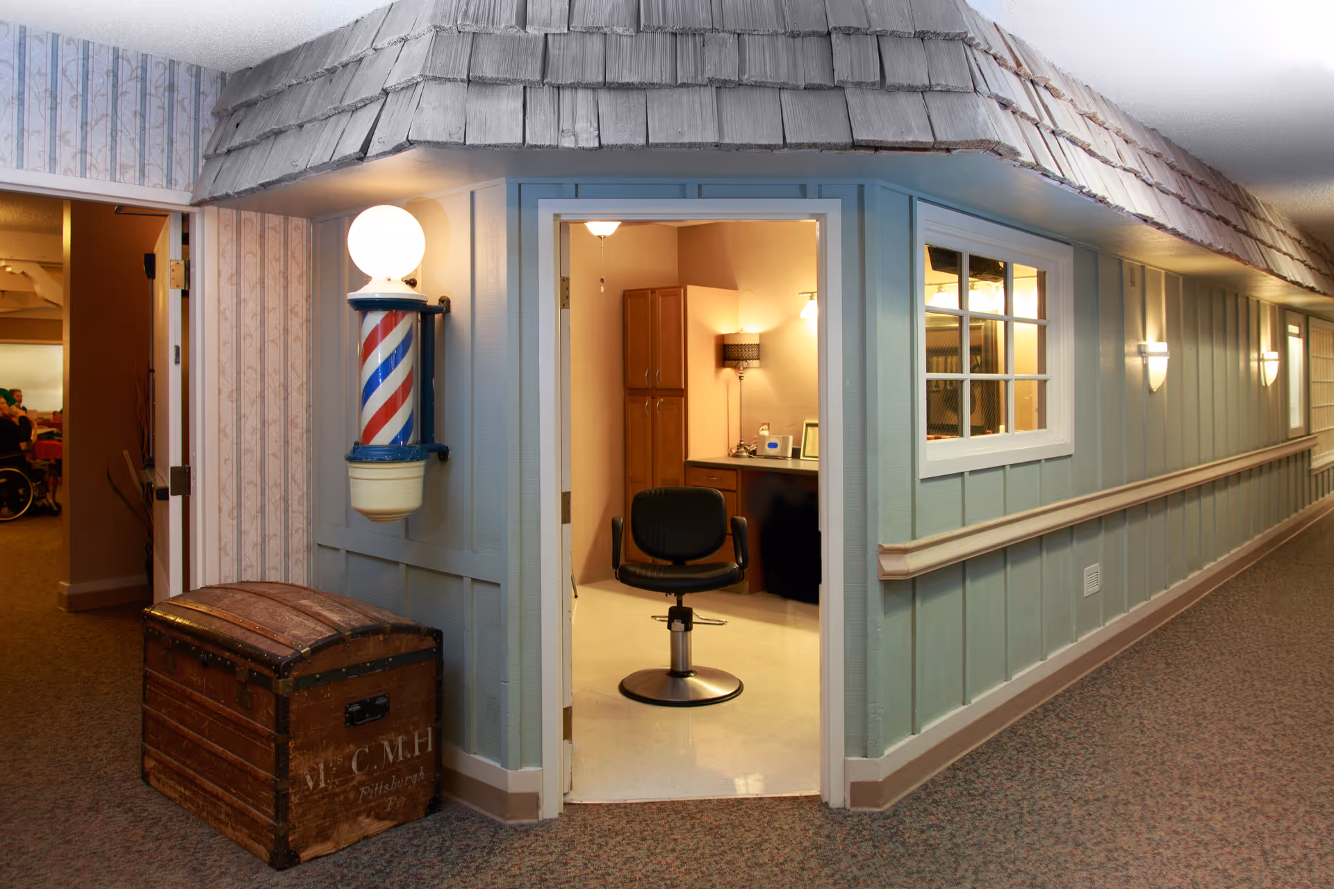 Indoor hallway of a senior facility with a small barber shop visible through an open door, a barber chair and a striped barber pole.