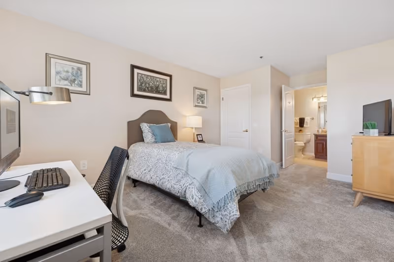 A neatly arranged bedroom with a single bed covered in a patterned bedspread and a light blue throw blanket. The room features a desk with a computer, keyboard, and mouse on the left side, a black mesh chair, and a wooden dresser with a TV on top on the right. The walls are decorated with three framed pictures. An open door reveals a bathroom with a toilet, sink, and mirror. The room has beige walls and carpeted flooring.