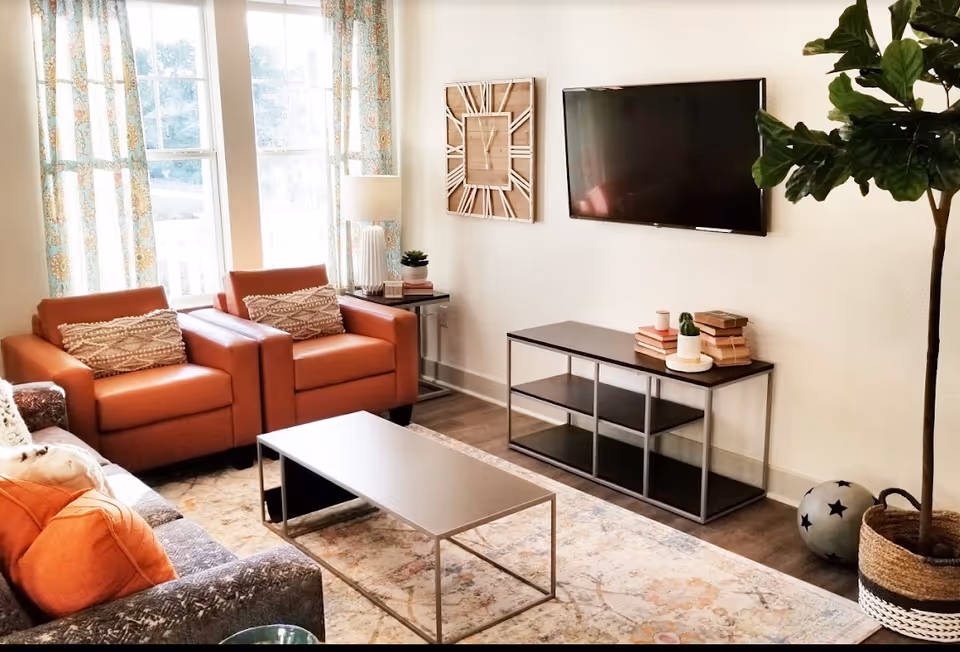 A bright living room with two orange armchairs, a gray sofa with orange pillows, a rectangular coffee table, a wall-mounted flat screen TV, a console table with books and small plants, a large decorative wall clock, patterned curtains on windows, and a tall potted plant in the corner.