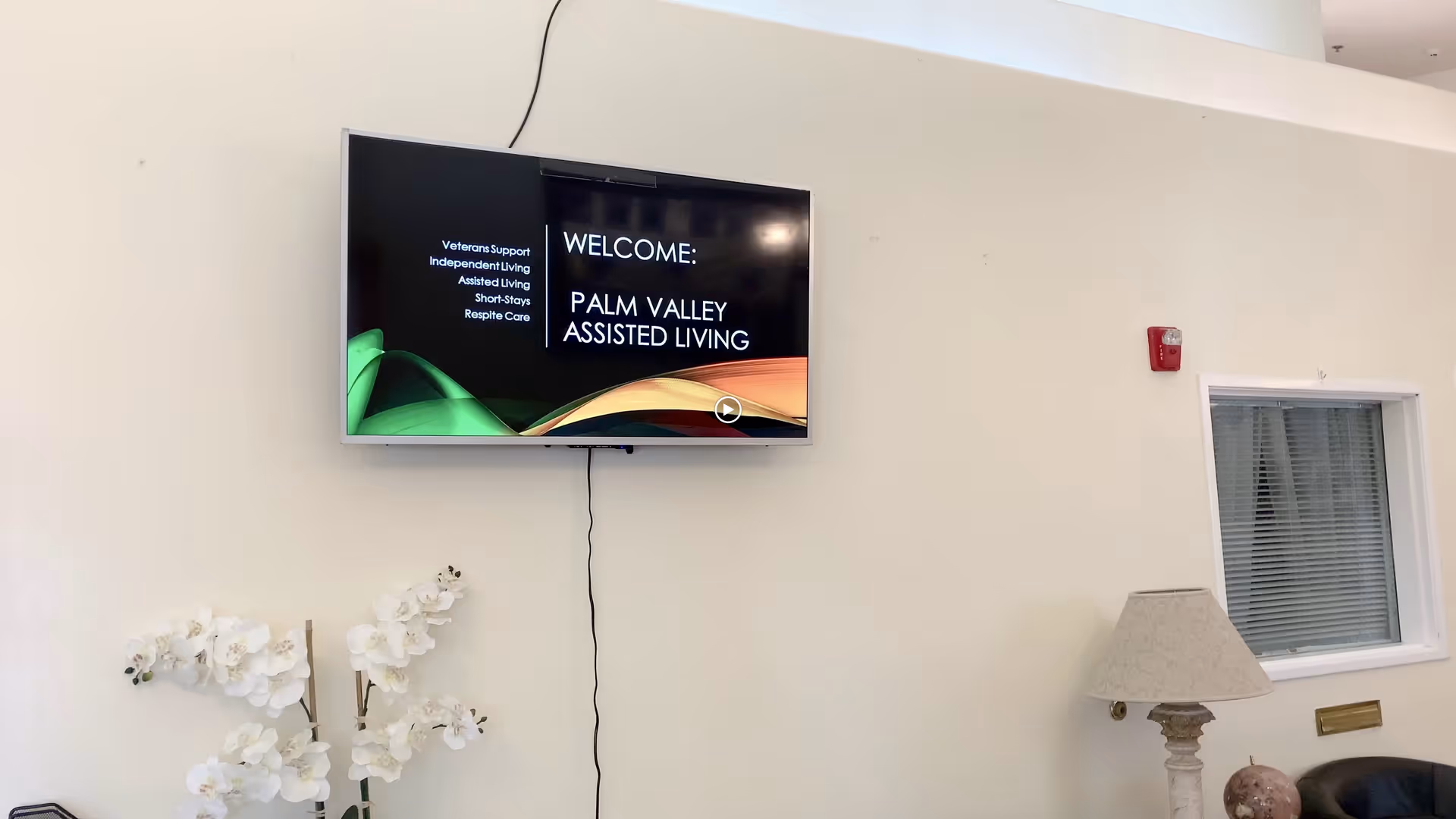 Interior wall of an assisted living facility with a mounted flat screen TV displaying a welcome message for Palm Valley Assisted Living. Below the TV is a white orchid plant. To the right, there is a table lamp on a small table next to a window with blinds and a fire alarm on the wall.
