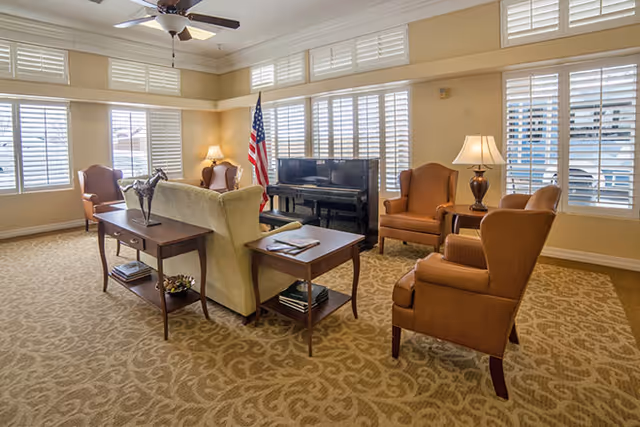A bright and spacious living room with beige patterned carpet, multiple armchairs, a sofa, side tables with lamps, a piano against the wall, and an American flag in the corner. Large windows with white shutters allow natural light to fill the room.