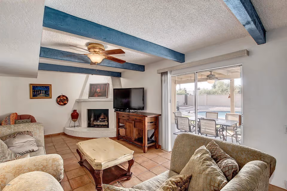 A cozy living room with beige sofas and patterned cushions, a wooden coffee table, a TV on a wooden stand, a white corner fireplace with decorative items, blue ceiling beams, a ceiling fan, and a large sliding glass door leading to an outdoor patio with a table, chairs, and a swimming pool.
