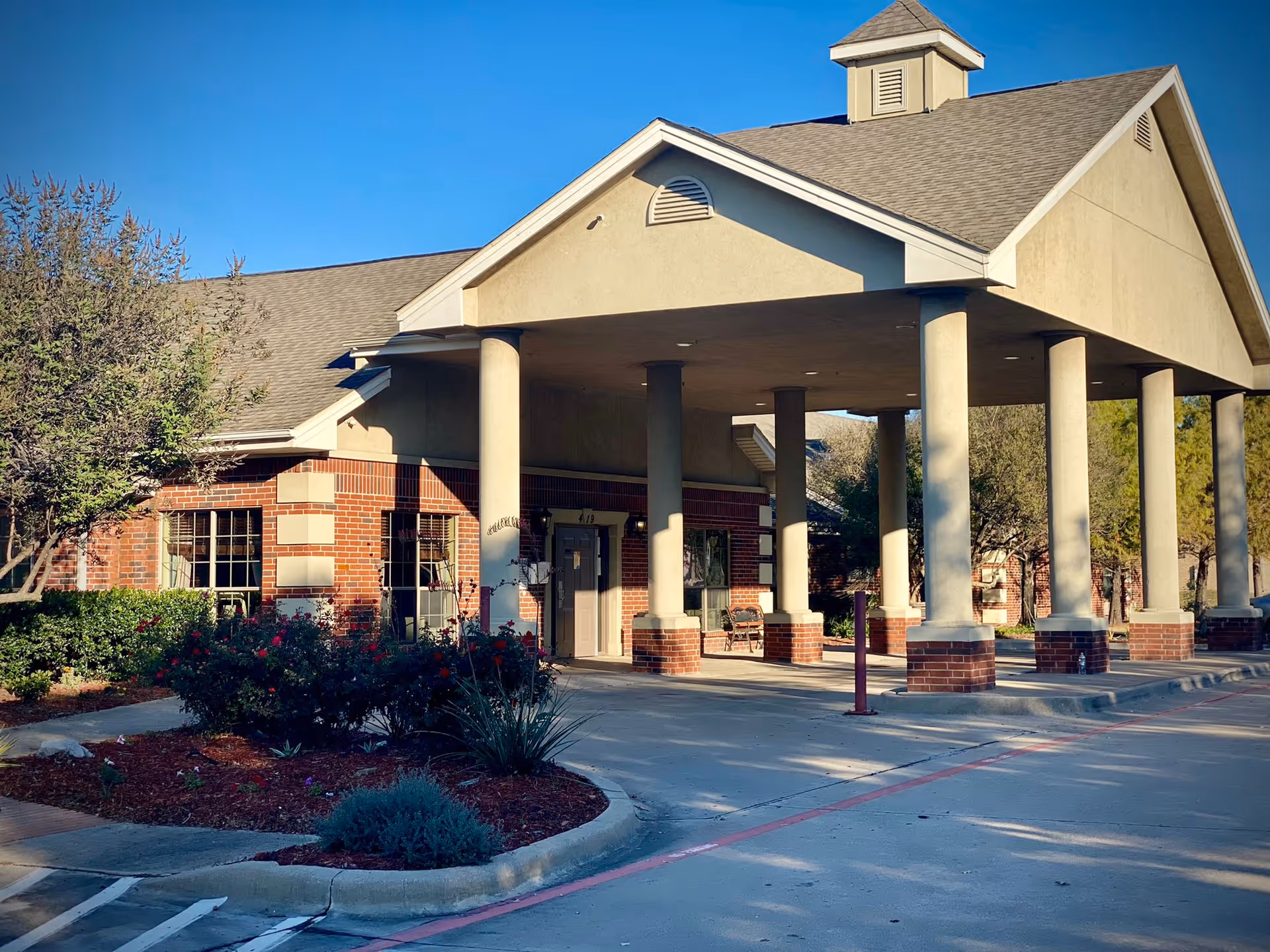 Front entrance of a brick and stucco building with a covered porte-cochère, columns, and landscaped beds.