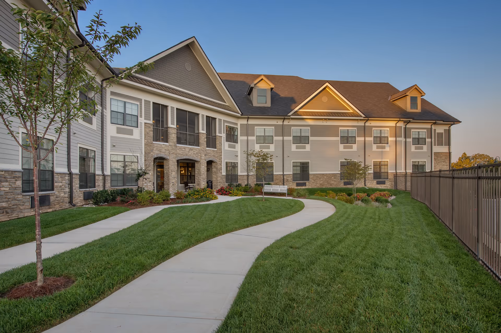 Exterior view of a two-story senior living facility building with beige and gray siding, stone accents, and multiple windows. A curved concrete pathway runs through a well-maintained green lawn with small trees and shrubs. A white bench is placed near the building, and a black metal fence borders the right side of the lawn under a clear blue sky.