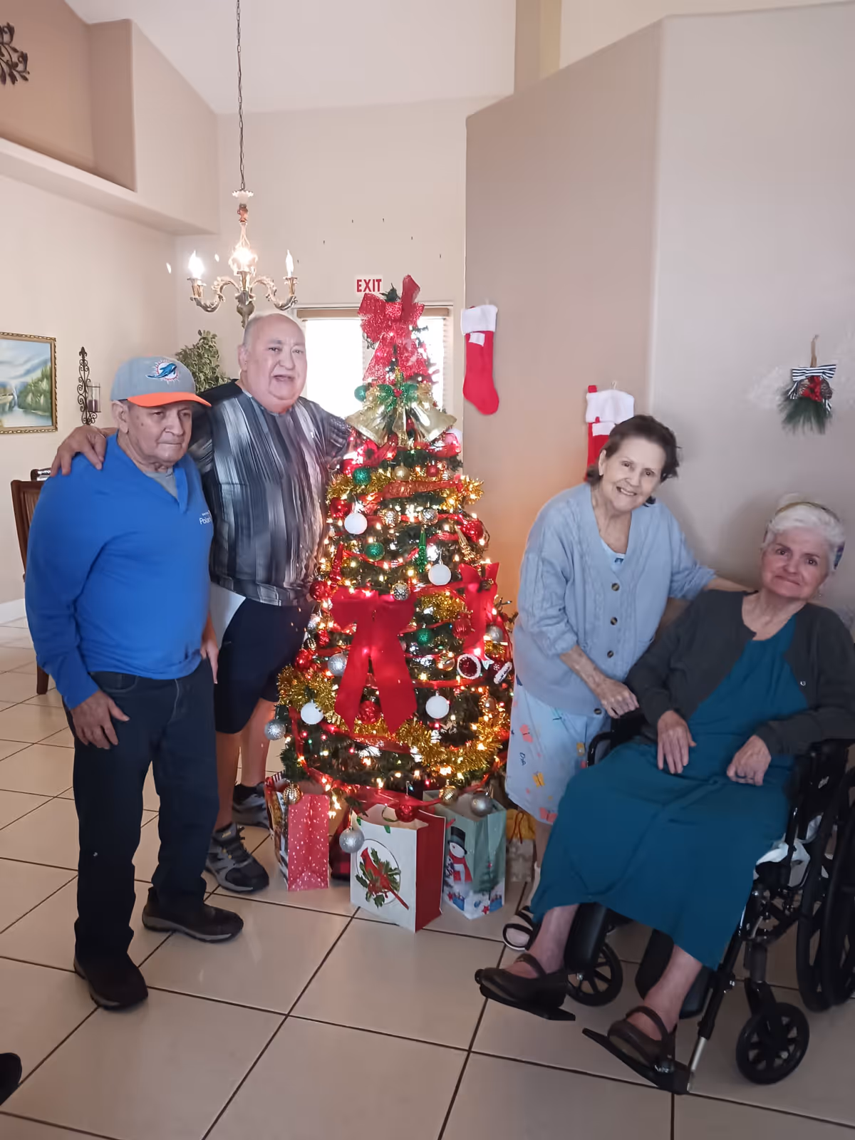 Four elderly people gathered around a decorated Christmas tree with presents underneath in a living room. Two men stand on the left side of the tree, one wearing a blue shirt and cap, the other in a striped shirt. On the right side, a woman in a light blue cardigan stands next to another woman seated in a wheelchair wearing a teal dress. The room has beige walls, a chandelier, and Christmas stockings hanging on the wall.