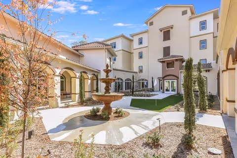 A beautifully landscaped courtyard featuring a central fountain, surrounded by greenery and the exterior of a multi-story building.