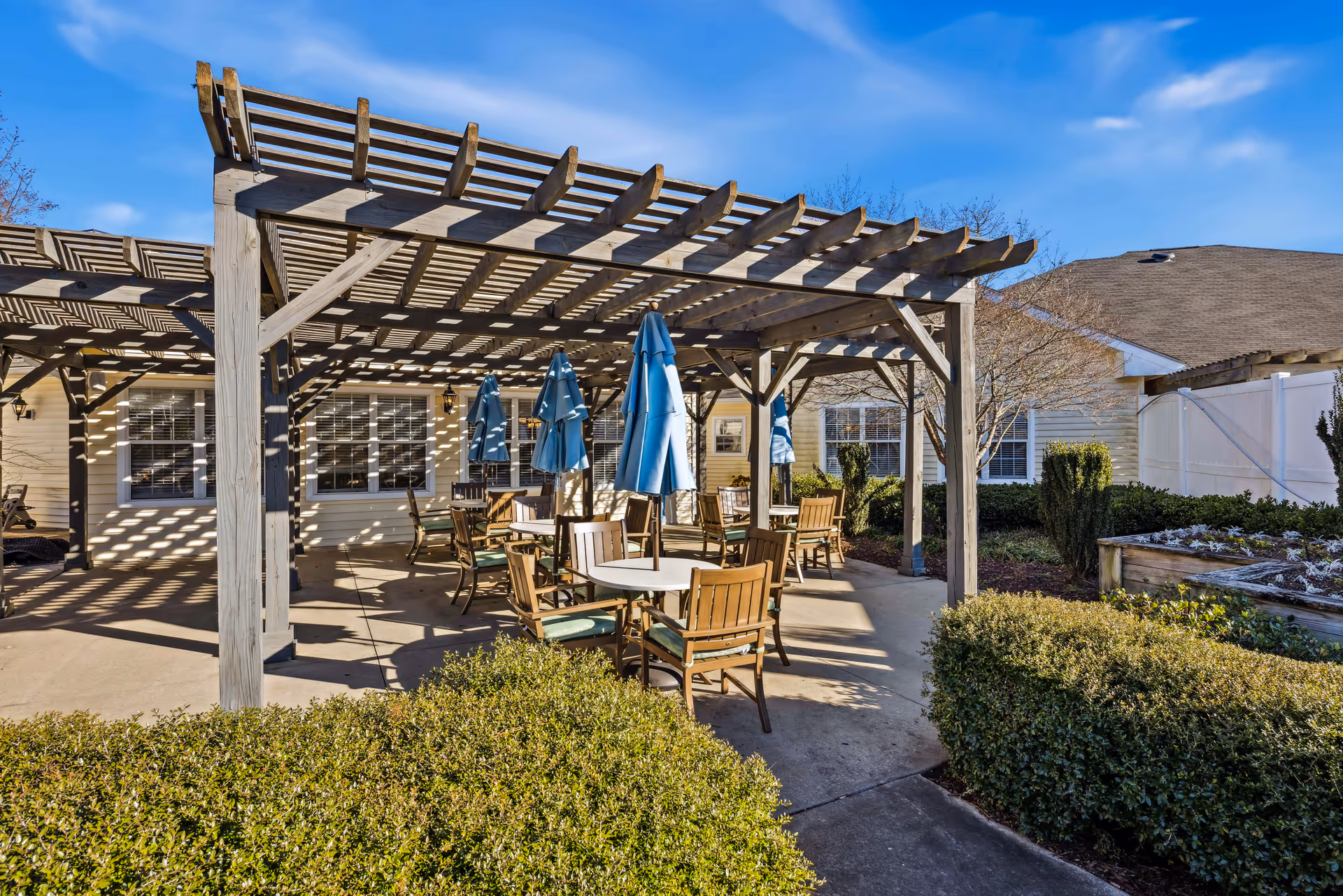 Outdoor patio area at TerraBella Fuquay-Varina with wooden pergolas providing partial shade over round tables and wooden chairs. Blue umbrellas are closed and positioned at the center of the tables. Surrounding the patio are bushes and a garden bed, with a building featuring windows in the background under a clear blue sky.
