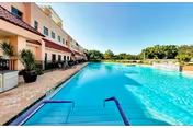 Outdoor swimming pool with clear blue water next to a multi-story building with balconies and patio seating. The pool area is surrounded by a paved deck with potted plants and greenery in the background under a clear blue sky.