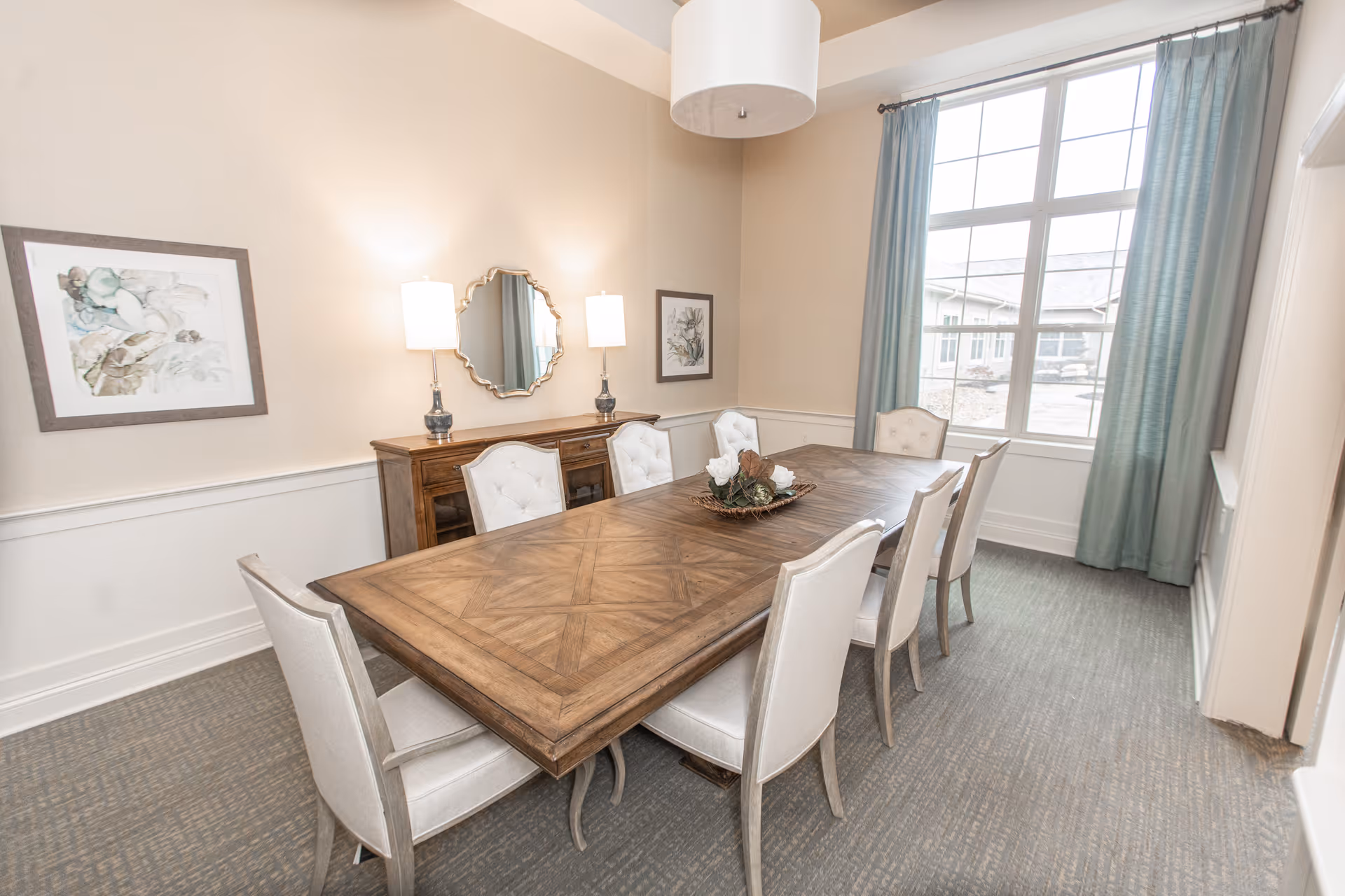 A bright dining room with a large wooden table surrounded by eight upholstered chairs. The room features beige walls with white wainscoting, two framed floral artworks, a decorative mirror, two table lamps on a wooden sideboard, and a large window with blue curtains letting in natural light.
