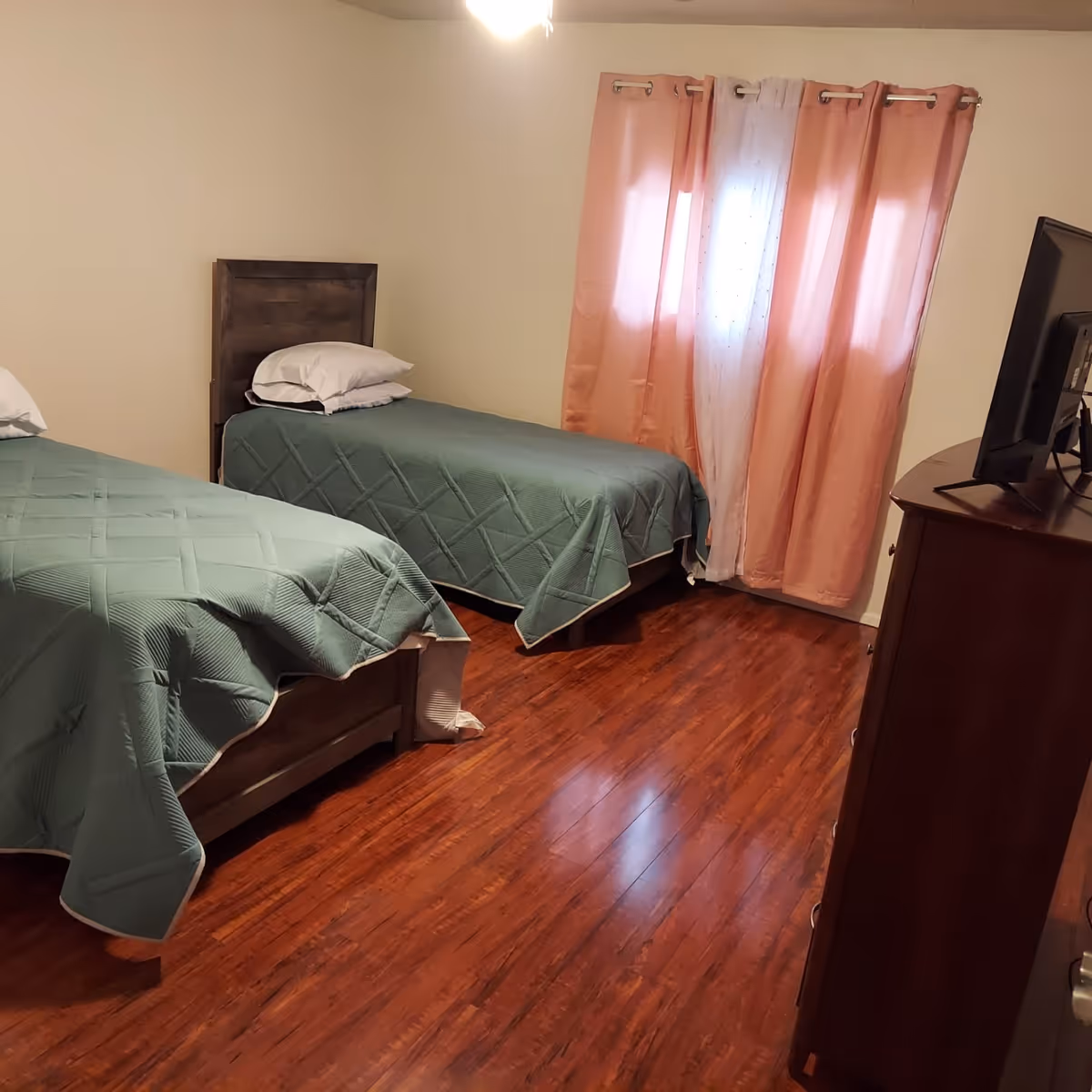 A bedroom with two single beds covered with green quilts, each with a pillow. The room has wooden flooring, a window with pink and white curtains, and a wooden dresser with a TV on top.