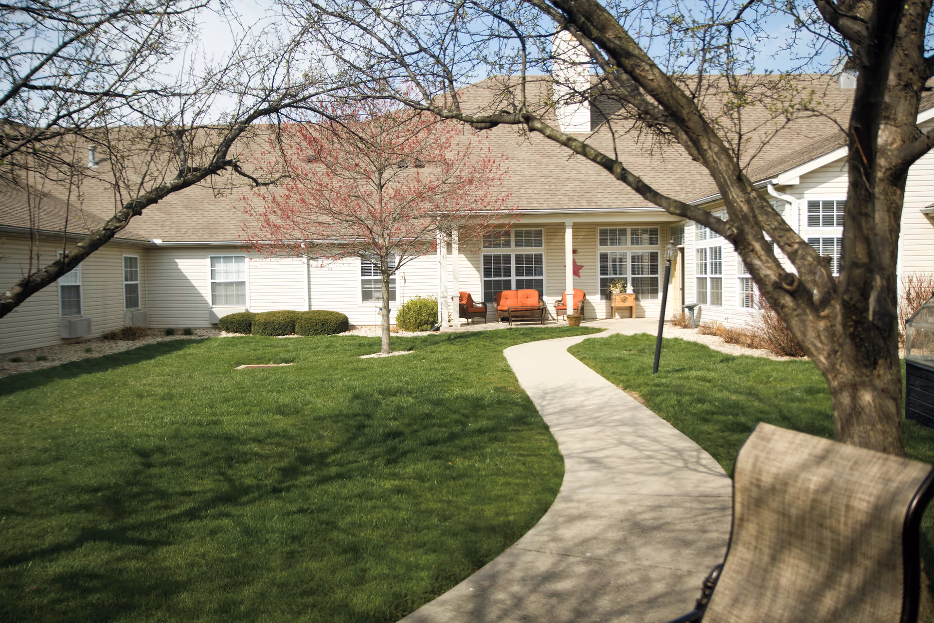 Curved concrete path through a grassy courtyard leading to the exterior entrance and patio of a single-story senior living building framed by trees.