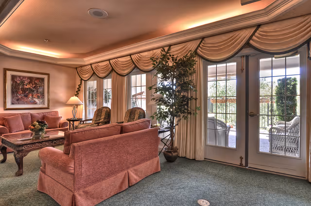 A cozy living room with pink upholstered sofas and armchairs arranged around a wooden coffee table. The room features large windows and glass doors with elegant draped curtains, allowing natural light to fill the space. A potted plant is placed near the doors, and a framed painting hangs on the wall above a side table with a lamp.