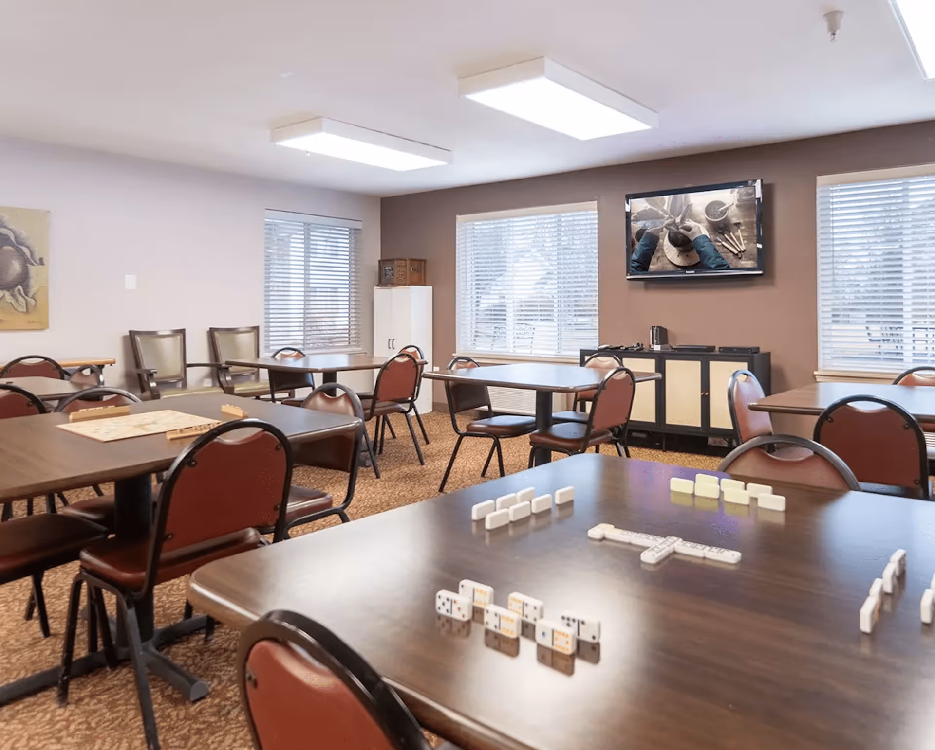 A well-lit common room with several tables and chairs arranged for group activities. Dominoes and a board game are set up on the tables. The room has large windows with blinds, a cabinet, and a wall-mounted TV displaying a pottery-making scene.