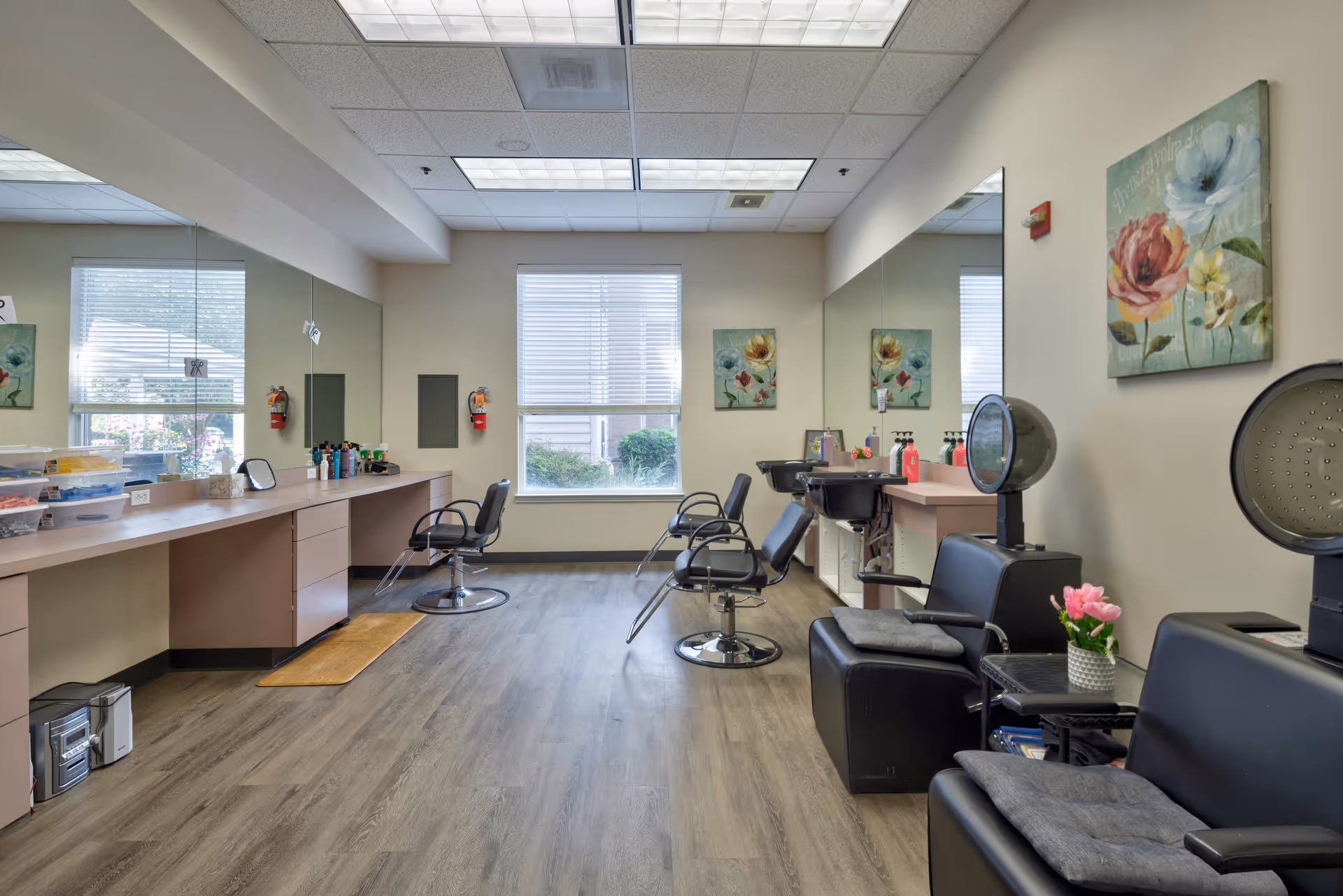 Interior view of a salon area in a senior living facility with salon chairs, hair washing sinks, large mirrors, and a window letting in natural light. The room has wood flooring, floral wall art, and a small table with a flower vase.
