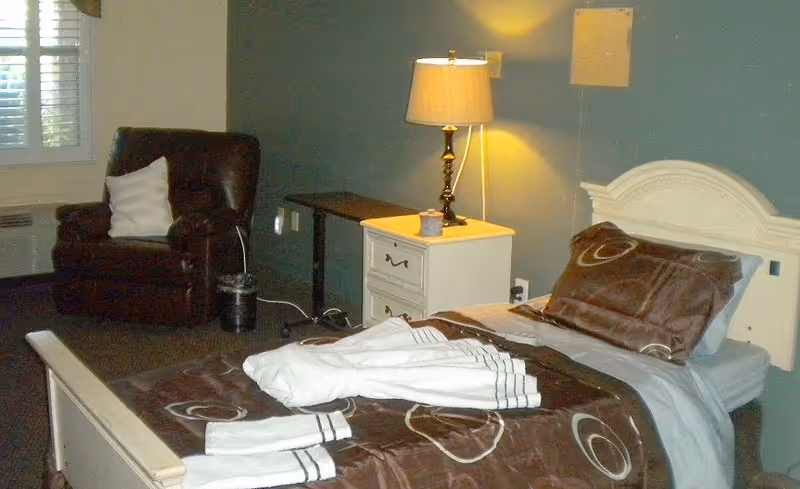 A single bed with brown and beige patterned bedding and a white towel folded on top, next to a white nightstand with a lamp. A dark brown armchair with a white pillow is positioned near a window with blinds in the background.