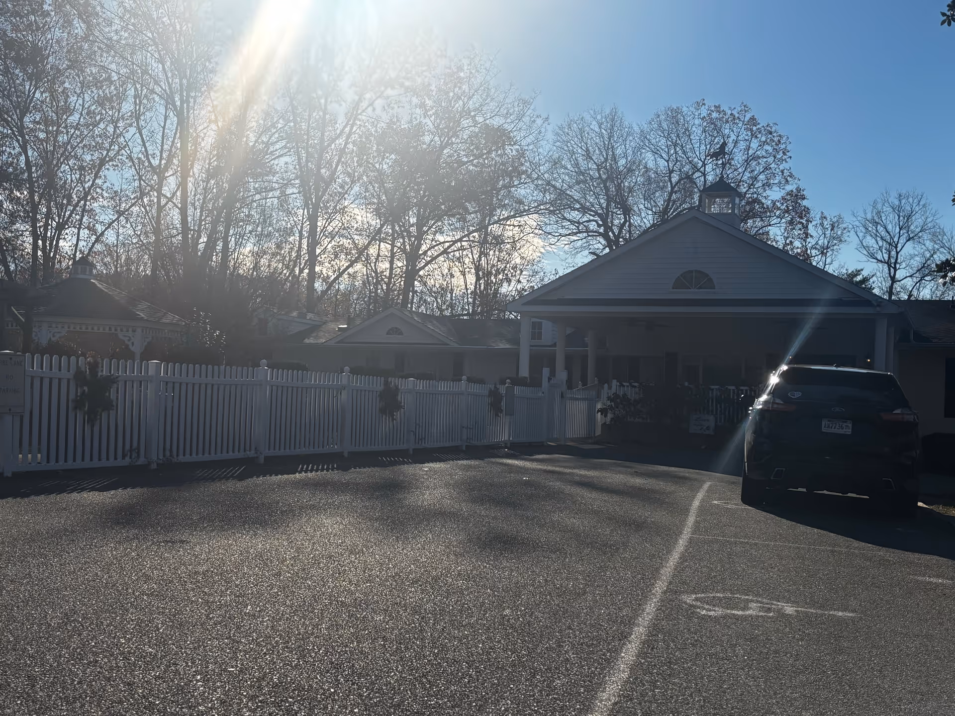 Front exterior of a single-story building with a white picket fence, a parked car in the lot, and bare trees with bright sun glare overhead.