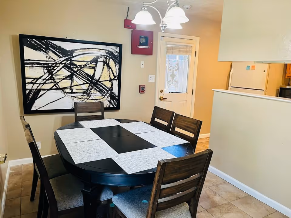 Dining area with a round black table covered with white placemats and surrounded by five wooden chairs with gray cushions. A large abstract black and white painting hangs on the beige wall. A white door with a window and a decorative metal grill is visible in the background, along with part of a kitchen area including a refrigerator and microwave. A ceiling light fixture with three lamps hangs above the table.