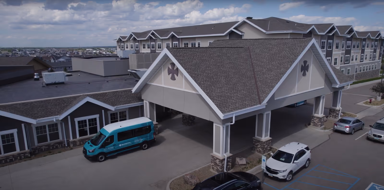 Exterior view of Benedictine Living Community-Bismarck showing a large covered entrance with a peaked roof supported by columns. Several cars and a teal shuttle van are parked in front of the building. The building has multiple stories with many windows and a cloudy sky in the background.
