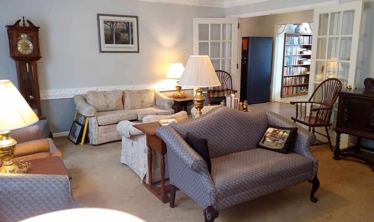 A cozy living room with a mix of vintage and classic furniture including a grandfather clock, two sofas, wooden chairs, side tables with lamps, and a bookshelf visible through glass-paneled double doors. The walls are painted light gray with white trim, and a framed picture hangs above one sofa.