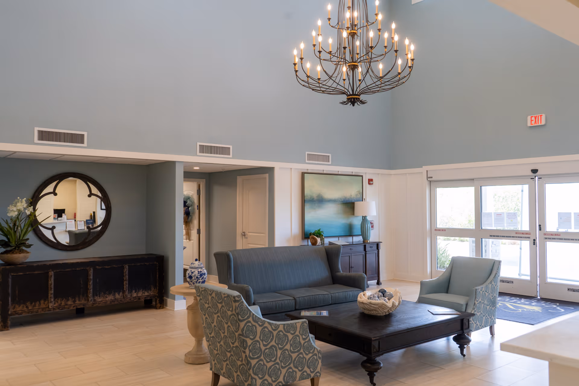 A bright and spacious lobby area with high ceilings and a large chandelier. The seating area includes a blue sofa and two patterned armchairs around a dark wooden coffee table. There is a sideboard with a round mirror and a potted plant on the left, and a cabinet with a lamp and artwork on the back wall near the entrance doors.