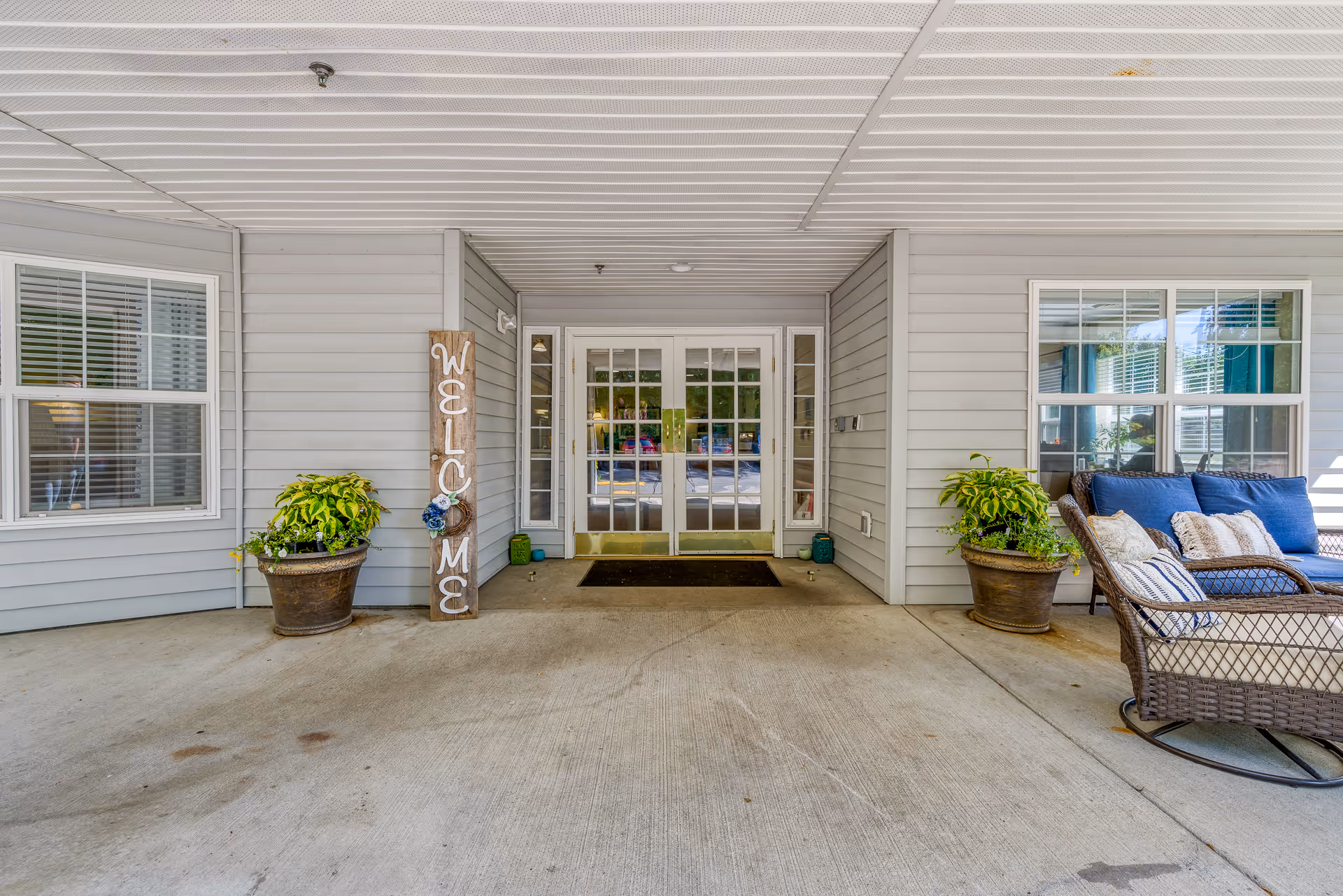 Covered front entrance with double glass doors, potted plants, a vertical 'WELCOME' sign, and wicker seating.