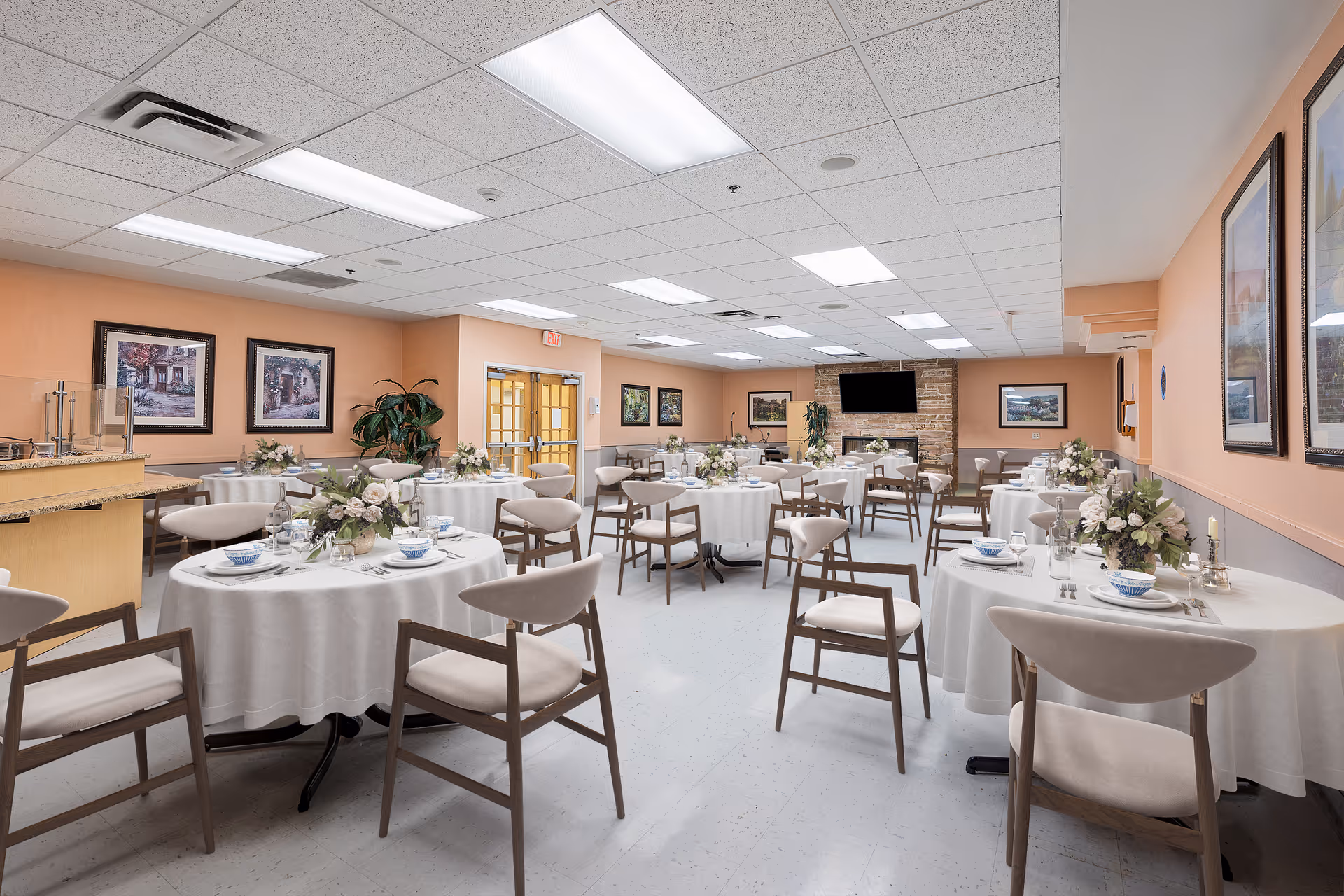 A spacious dining room with round tables covered in white tablecloths, each set with plates, bowls, glasses, and floral centerpieces. The room has peach-colored walls adorned with framed artwork, a stone fireplace with a mounted TV above it, and a ceiling with fluorescent lighting. Several wooden chairs with cushioned seats are arranged around the tables.