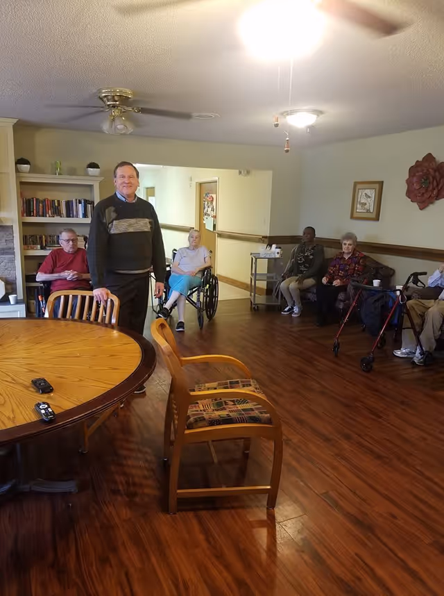 A group of elderly people sitting and standing in a senior living facility common area with wooden floors, a round wooden table with chairs, a bookshelf, and wall decorations. One man is standing near the table, while others are seated in chairs, a wheelchair, and using a walker.
