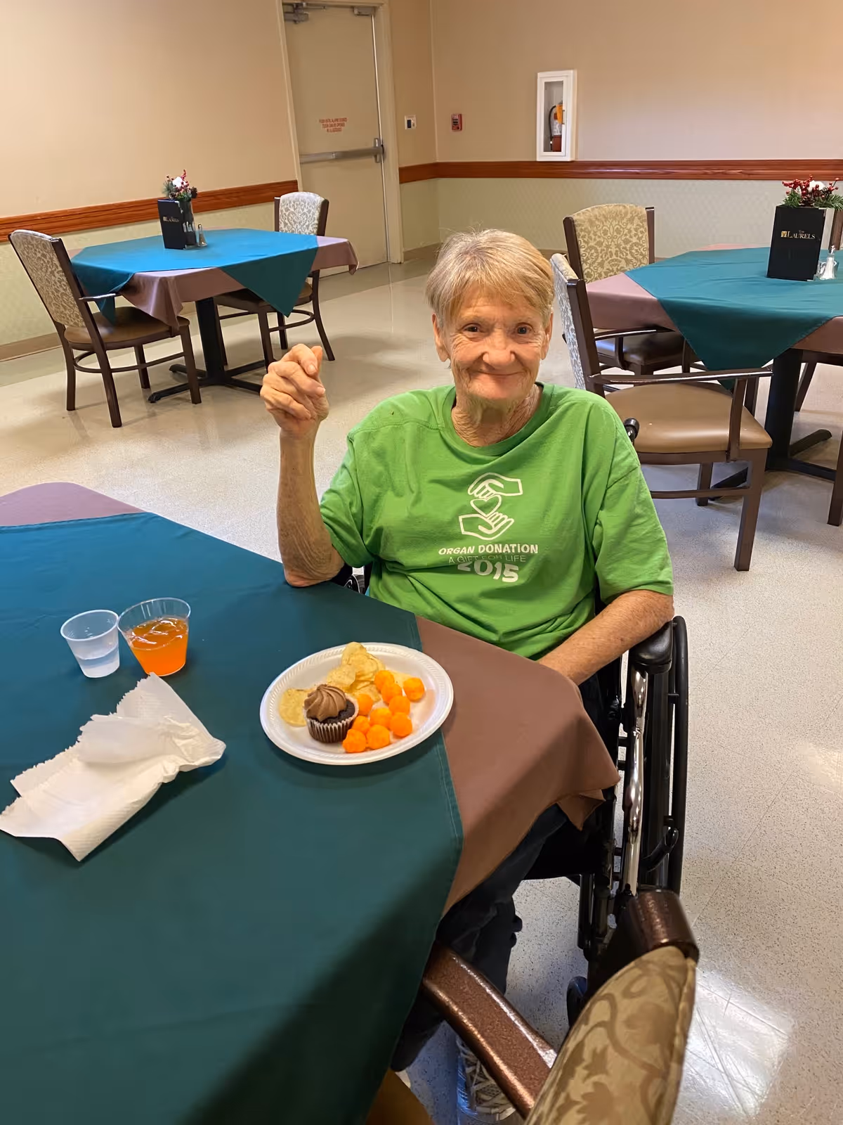 An elderly woman in a wheelchair sitting at a table in a dining area. She is wearing a green t-shirt and smiling at the camera. On the table in front of her is a plate with a chocolate cupcake, potato chips, and cheese balls, along with two cups containing water and an orange beverage. The room has several tables with green and brown tablecloths and chairs, and a beige wall with a fire extinguisher cabinet is visible in the background.