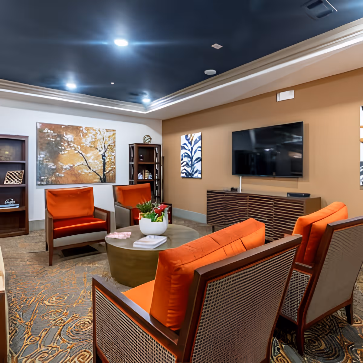 A cozy living room with orange cushioned chairs arranged around a round coffee table with a plant and books on it. The room features a flat-screen TV mounted on a beige wall above a wooden cabinet, decorative paintings on the walls, and a bookshelf in the corner. The ceiling has recessed lighting and crown molding.