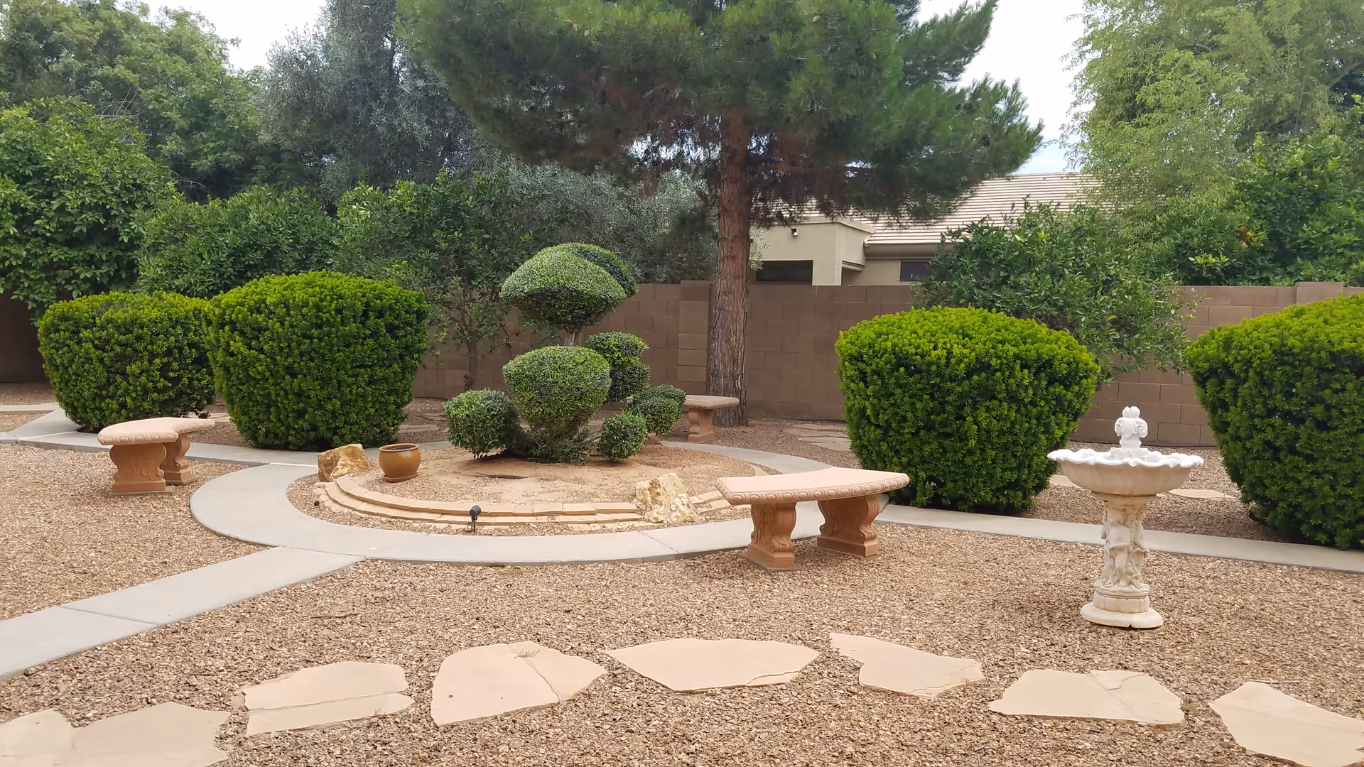 A landscaped outdoor garden area with neatly trimmed bushes, a small tree, stone benches, a birdbath, and a circular stone feature surrounded by gravel and paved walkways. A brick wall and some trees are visible in the background.