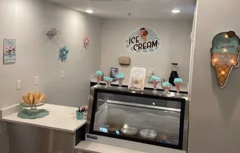 Interior view of an ice cream serving area in a senior living facility with a display freezer containing ice cream containers, several ice cream sundaes on top, and ice cream-themed wall decorations including a sign that says 'ICE CREAM' and a lighted ice cream cone sign.