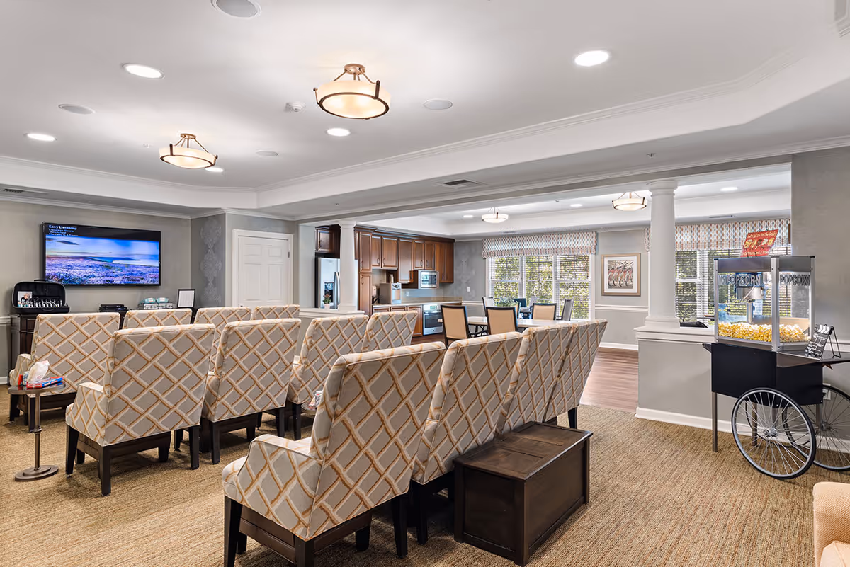 A cozy common area with rows of patterned upholstered chairs facing a wall-mounted flat screen TV. To the right, there is a popcorn machine on a cart. The room has beige carpeting, soft lighting from ceiling fixtures, and large windows with patterned valances letting in natural light. In the background, there is a dining area with a table and chairs and a kitchen with wooden cabinets.