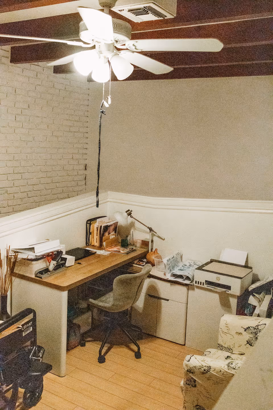 A small office corner with a wooden desk cluttered with papers, books, and office supplies. There is a gray swivel chair in front of the desk, a white printer on a cabinet to the right, and a floral-patterned armchair partially visible. The room has light-colored walls with a brick accent wall on the left and a ceiling fan with lights above.