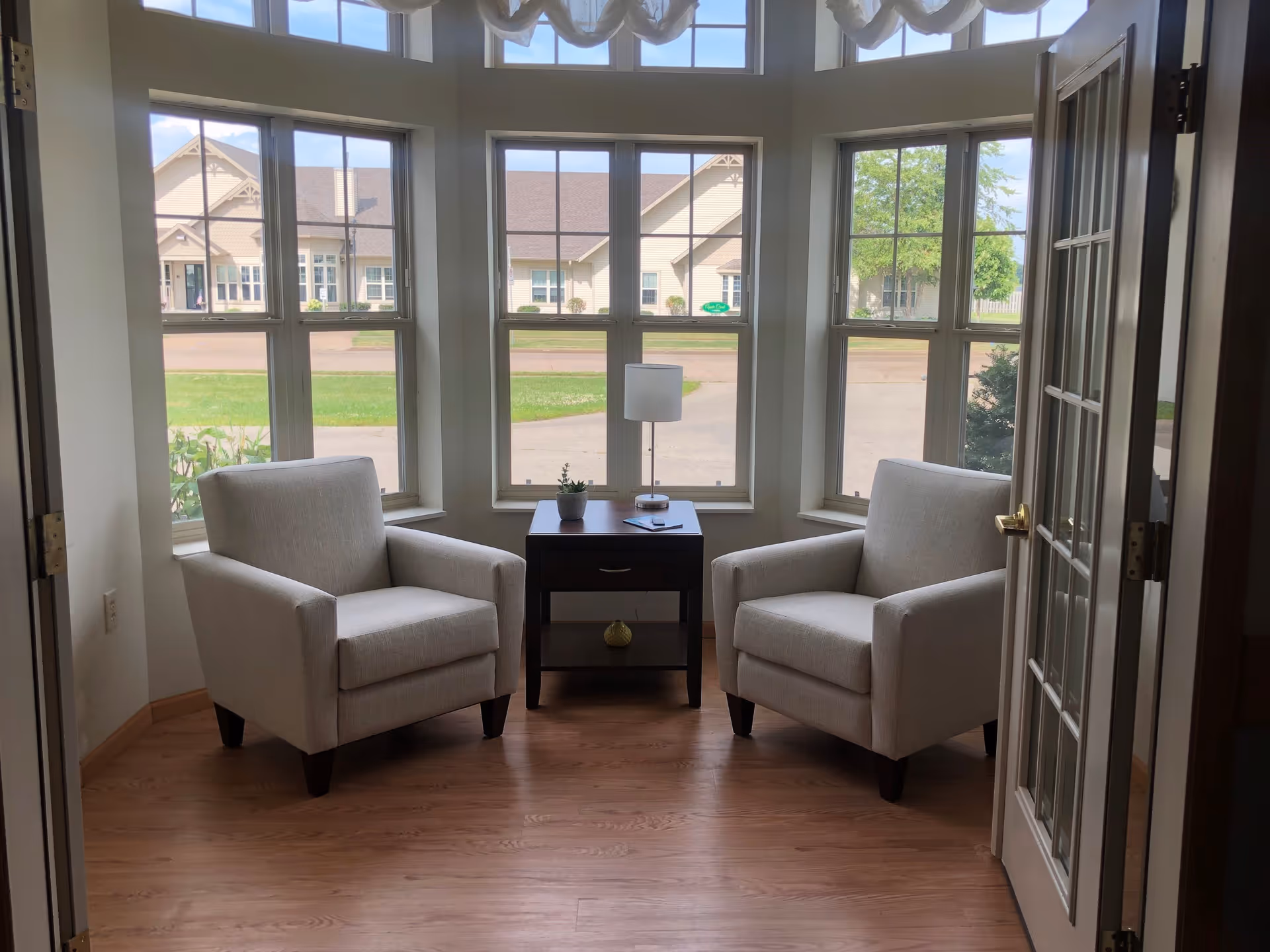 A cozy sitting area with two beige armchairs facing each other, separated by a small dark wooden table with a lamp and a small plant. The area is surrounded by large windows showing a view of a building and greenery outside. The floor is wooden, and there is a glass-paneled door partially open on the right side.