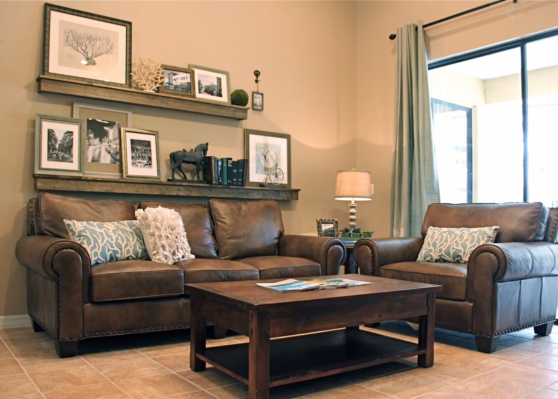 A cozy living room with a brown leather sofa and matching armchair, decorated with patterned and textured throw pillows. A wooden coffee table sits in front of the sofa with magazines on top. Behind the sofa, two wooden shelves display framed pictures, books, and decorative items. A table lamp and small plants are on a side table next to the armchair. Light-colored curtains frame a large window letting in natural light.