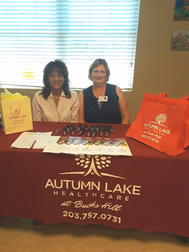 Two women sitting behind a table covered with a maroon tablecloth that has the Autumn Lake Healthcare at Bucks Hill logo and phone number. On the table are brochures, pens, and promotional bags in yellow and orange colors with the same logo and contact information. A window with blinds is in the background.