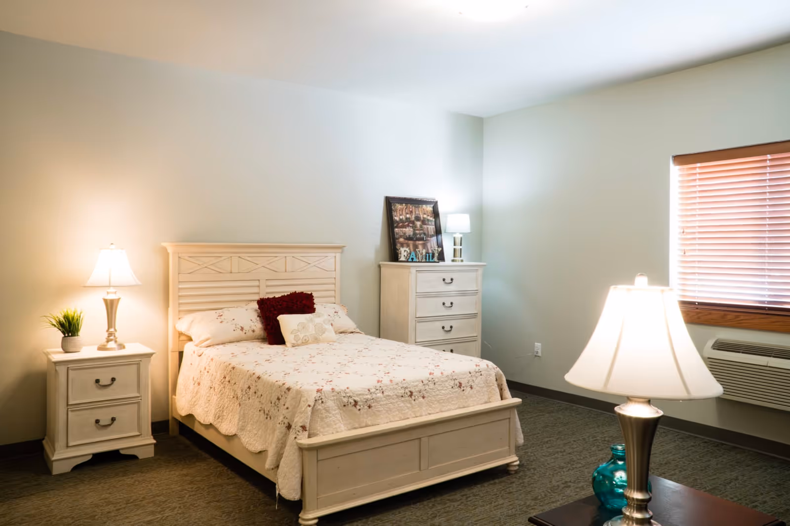 A cozy bedroom with a white wooden bed frame and floral bedspread. There are two bedside tables with lamps on each side of the bed, a chest of drawers with a lamp and decorative items, a window with wooden blinds, and a carpeted floor.