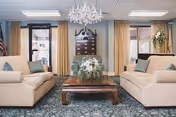 A cozy living room with two beige sofas facing each other across a wooden coffee table adorned with a floral arrangement. The room features a patterned carpet, large windows with beige curtains, and a decorative chandelier hanging from the ceiling. A dark wooden wall-mounted cabinet is centered between the windows.