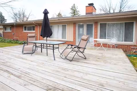 Back wooden patio with table, umbrella, and lounge chairs by a brick senior living facility.