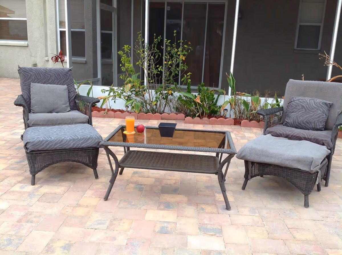 Outdoor patio area with two cushioned wicker chairs and matching ottomans facing a glass-top wicker coffee table. On the table, there is a glass of orange juice, a red apple, and a small black planter. Behind the seating area is a garden bed with various green plants and a building with large windows.