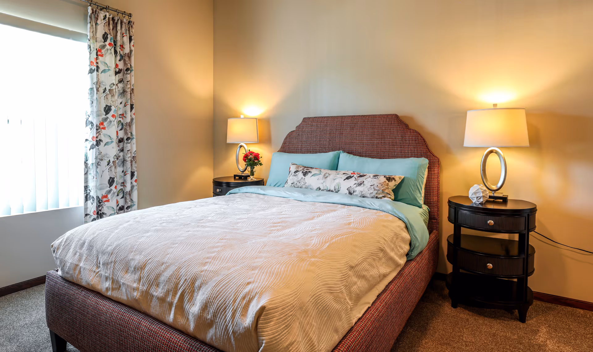 A cozy bedroom with a neatly made bed featuring a textured beige comforter, light blue pillows, and a floral accent pillow. The bed has a reddish-brown upholstered headboard. On each side of the bed are black nightstands with drawers, each holding a modern lamp with a circular base. A window with floral curtains allows natural light into the room.